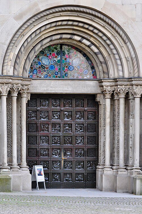 Porta principal da Grossmünster em Zurique Suíça mostrando portal românico com arcos concêntricos, vitral colorido de Augusto Giacometti em formato de luneta, portas de bronze com relevos e colunas decoradas