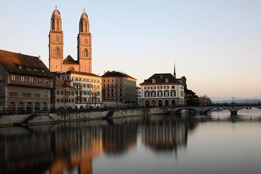 Grossmünster Zurique Suíça mostrando catedral com duas torres gêmeas refletidas no Rio Limmat ao amanhecer, edifícios históricos à margem do rio e ponte em dia calmo