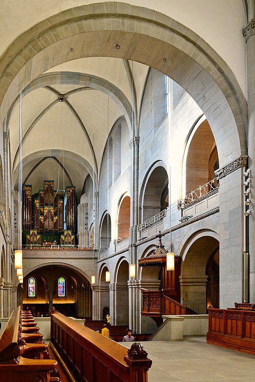 Interior da Grossmünster em Zurique Suíça mostrando nave central com bancos de madeira, órgão de tubos ao fundo na tribuna, púlpito de madeira escura ornamentado, arcos românicos em pedra clara e vitrais coloridos