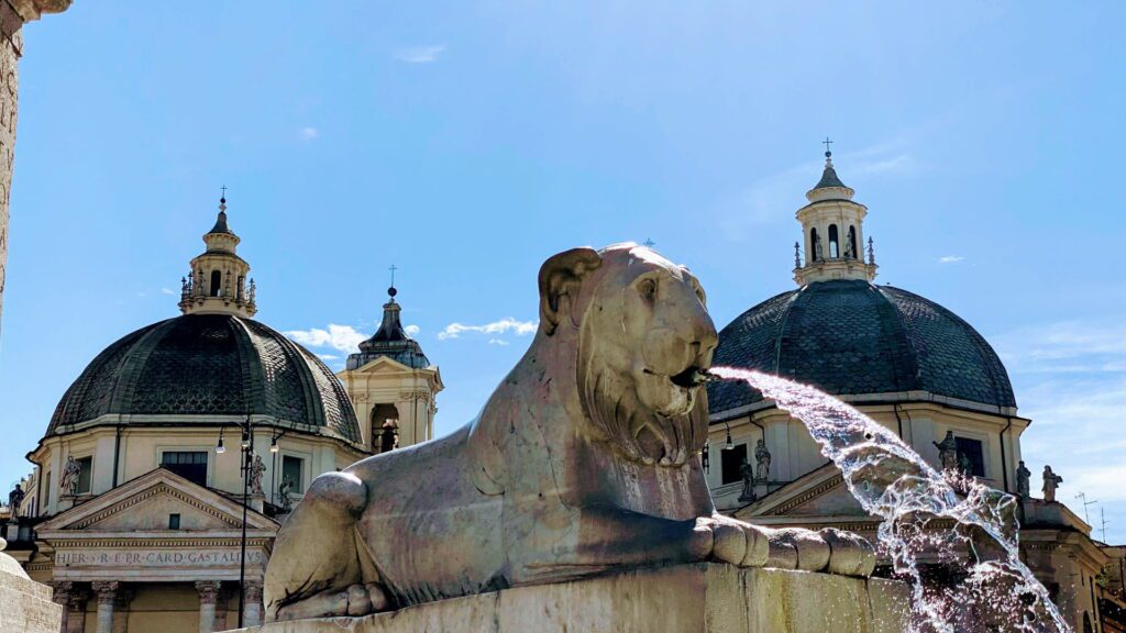 Leão de pedra cuspindo água na fonte da Piazza del Popolo com cúpulas das igrejas gêmeas Santa Maria dei Miracoli e Santa Maria in Montesanto ao fundo, Roma