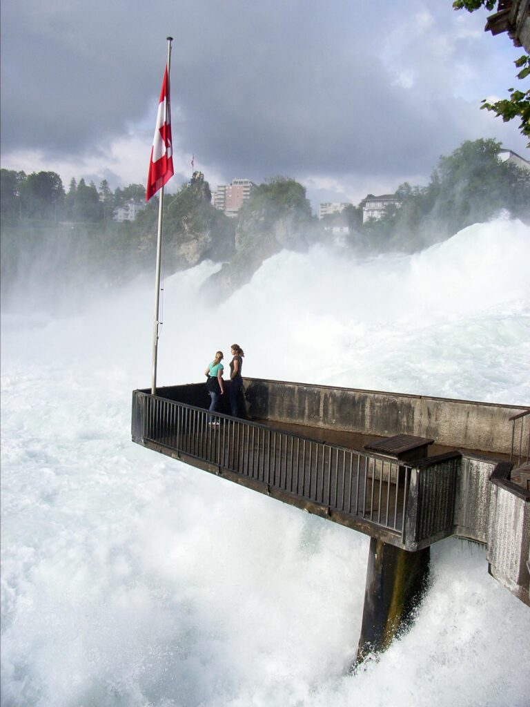 Mirante sobre as Cataratas do Reno na Suíça com bandeira suíça vermelha hasteada, duas pessoas observando a cachoeira poderosa de plataforma de madeira, névoa intensa das águas e edifícios ao fundo sob céu nublado