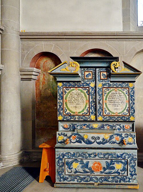 Móvel histórico decorado com pinturas de arte popular tradicional mostrando painéis com inscrições em molduras ovais, ornamentos florais em azul amarelo e laranja, volutas douradas e motivos decorativos barrocos em igreja