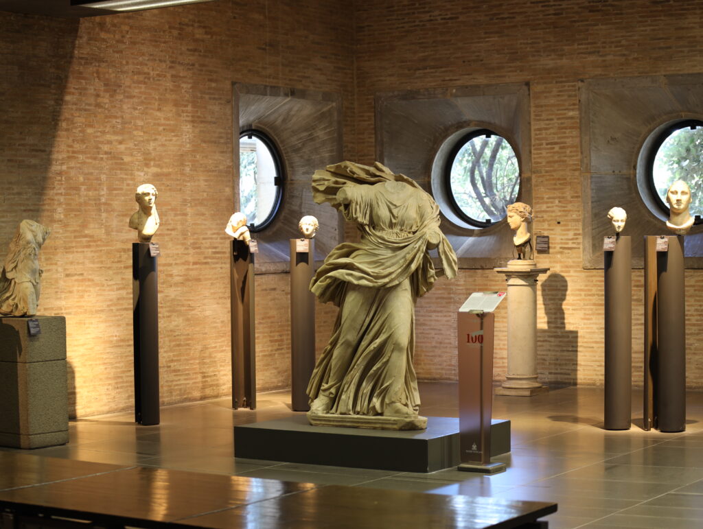 Sala do Museo Gregoriano Profano com escultura de Nike/Vitória e bustos romanos, arquitetura com paredes de tijolos e janelas circulares, Vaticano