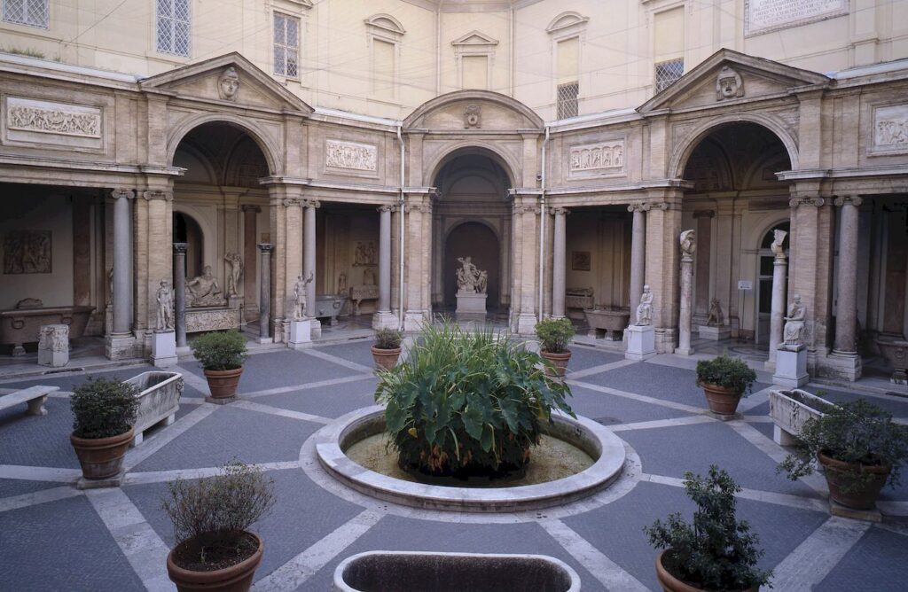 Pátio aberto dos Museus do Vaticano com arquitetura clássica, arcos, colunas e turistas durante a visita ao complexo.