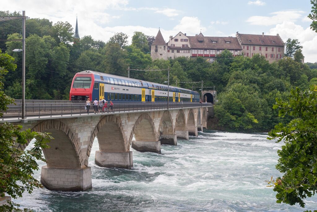 Trem SBB vermelho e azul cruzando ponte de arcos de pedra sobre o Rio Reno na Suíça, com águas turbulentas abaixo, Castelo de Laufen medieval no topo da colina arborizada e túnel ferroviário ao fundo