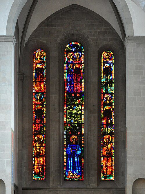 Três vitrais coloridos altos e estreitos na Grossmünster Zurique mostrando figuras religiosas estilizadas em tons vibrantes de azul, vermelho, laranja e verde, inseridos em arcos góticos de pedra cinza