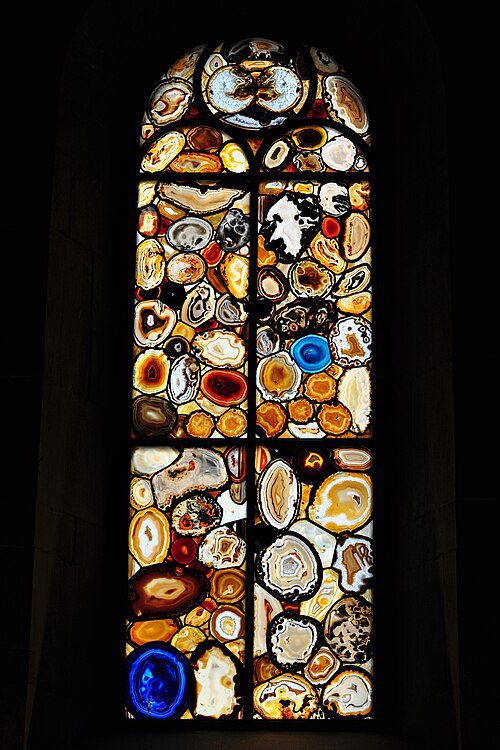Vitral artístico de Augusto Giacometti na Grossmünster Zurique com formas orgânicas coloridas lembrando seções de ágata e pedras preciosas em tons de âmbar, branco e azul intenso, iluminado por luz natural