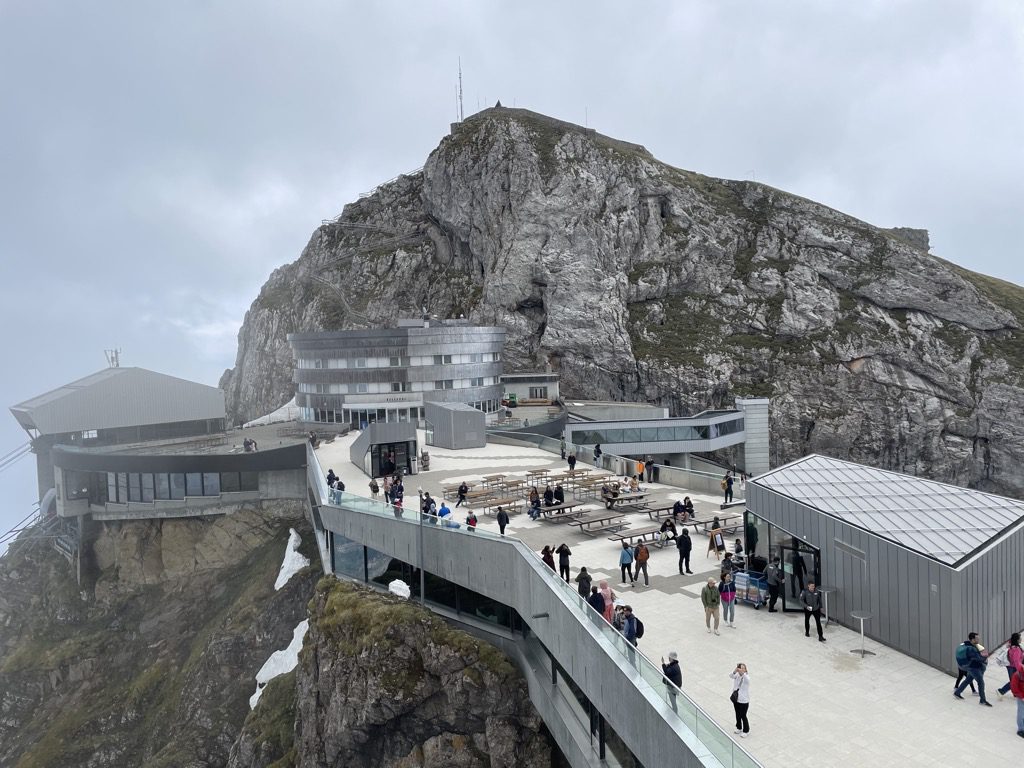 Vista da praça do cume do Monte Pilatus (Pilatus Kulm, 2.128 m): o edifício circular do hotel e restaurante, terraços com mesas e turistas, a passarela envidraçada da estação do teleférico e o pico rochoso com antenas acima, sob céu encoberto, Lucerna, Suíça