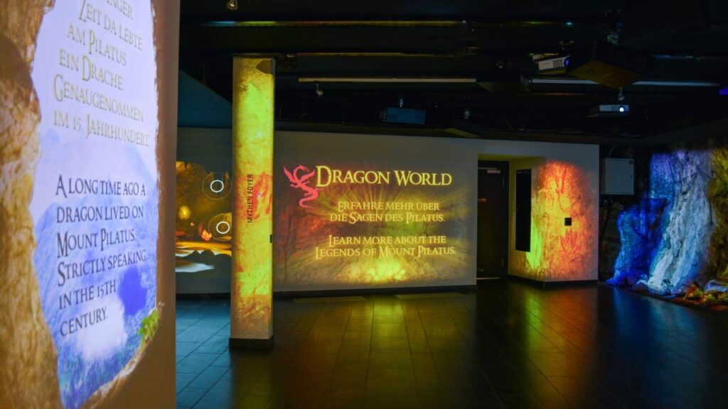 Interior da exposição interativa "Dragon World" no Pilatus Kulm: projeções coloridas nas paredes e colunas, painel com dragão vermelho e textos bilíngues sobre as lendas do Monte Pilatus, Lucerna, Suíça