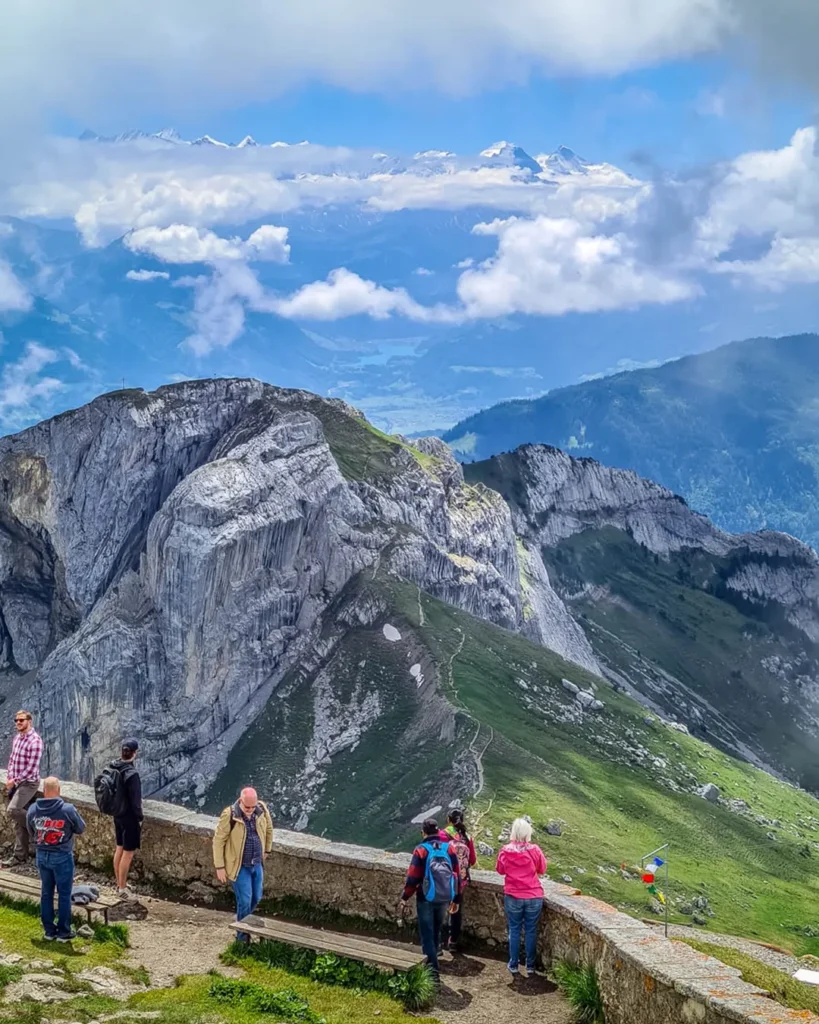 Turistas apoiados no muro de pedra do mirante do cume do Pilatus (Pilatus Kulm, 2.128 m) admiram os paredões de calcário do maciço e, ao fundo, os picos nevados dos Alpes Suíços emergindo de um mar de nuvens, Lucerna, Suíça