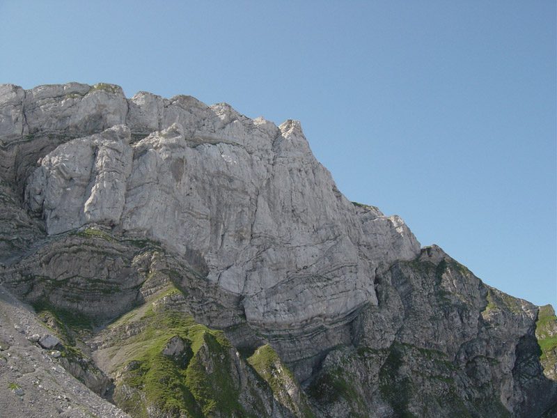 Close das formações de calcário alpino do Monte Pilatus: paredão claro com estratos sedimentares dobrados, encostas de grama e cascalho sob céu azul, na região de Lucerna, Suíça