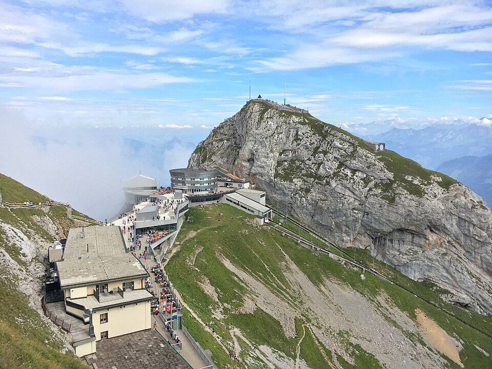 Vista do alto do Pilatus Kulm (2.128 m): hotel histórico, restaurante panorâmico e terraços com turistas, com o trilho da ferrovia de cremalheira mais íngreme do mundo descendo a encosta e os Alpes ao fundo, Lucerna, Suíça