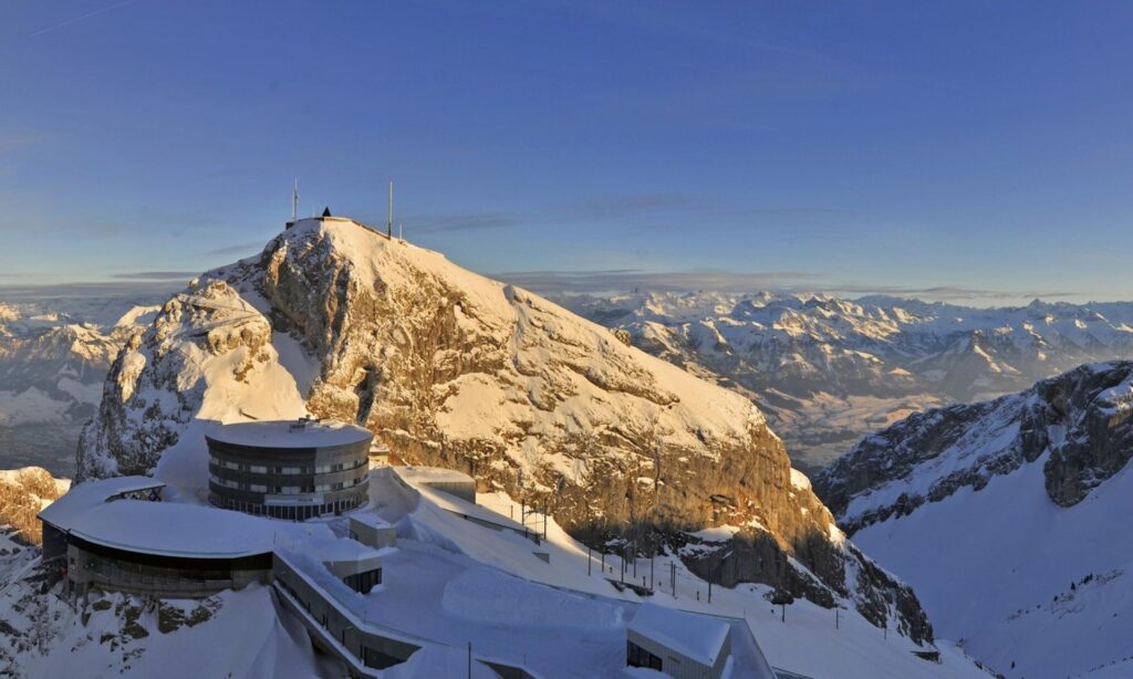Vista de inverno do cume do Monte Pilatus (Pilatus Kulm, 2.128 m): o hotel circular e as estruturas do cume cobertos de neve sob a luz dourada do entardecer, com o panorama dos Alpes Suíços nevados até o horizonte, Lucerna, Suíça