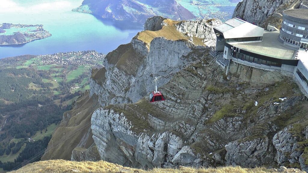 Vista do cume do Pilatus: cabine vermelha do teleférico Fräkmüntegg–Pilatus Kulm ("Dragon Ride") sobre os paredões de calcário, com a estação do cume à direita e o Lago de Lucerna turquesa lá embaixo, Suíça
