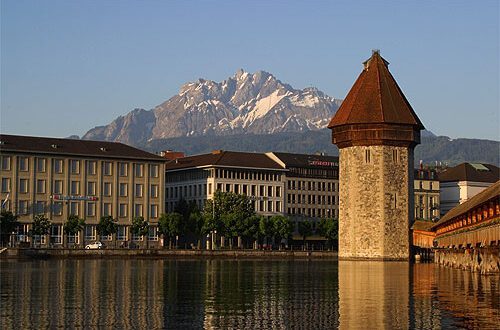 A Torre da Água banhada pela luz dourada, com um trecho da Ponte da Capela à direita, prédios da margem do rio Reuss à esquerda e o maciço nevado do Monte Pilatus ao fundo, em Lucerna, Suíça