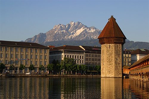 A Torre da Água banhada pela luz dourada, com um trecho da Ponte da Capela à direita, prédios da margem do rio Reuss à esquerda e o maciço nevado do Monte Pilatus ao fundo, em Lucerna, Suíça