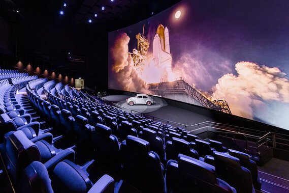 Teatro moderno com tela curva gigante exibindo lançamento de ônibus espacial, fileiras de poltronas azuis e um carro branco exposto na plataforma frontal.