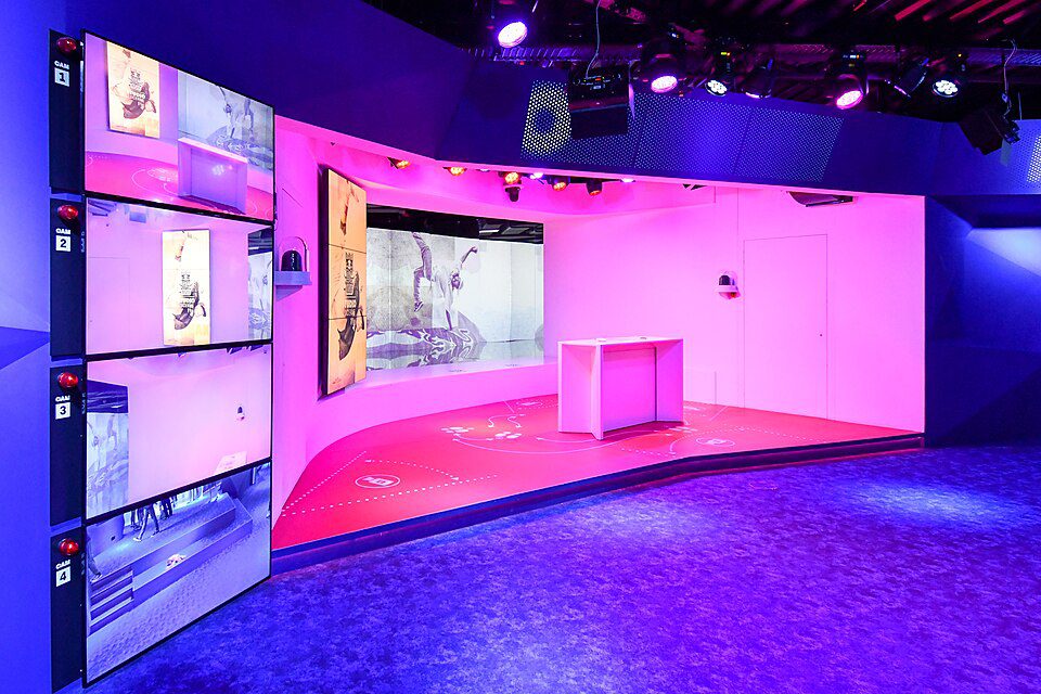Estúdio interativo com iluminação rosa e azul, parede de monitores empilhados, plataforma elevada com mesa branca e projeções, espaço tecnológico moderno