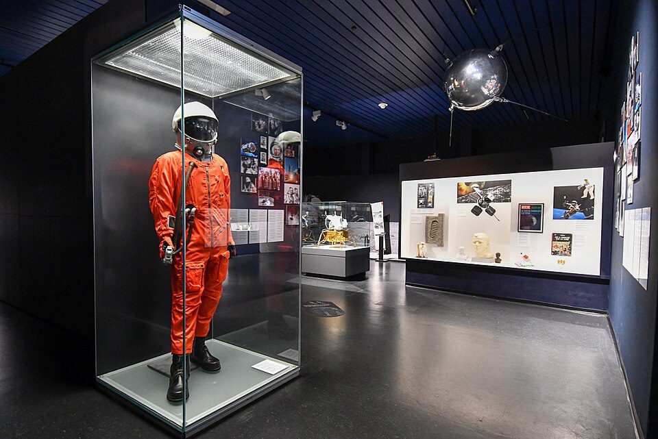 Exposição de museu espacial com traje de voo laranja em vitrine de vidro e réplica do satélite Sputnik pendurada no teto