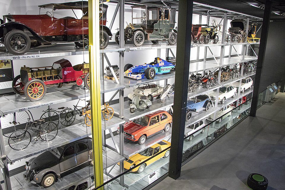 Estantes de museu organizadas em múltiplos níveis exibindo uma vasta coleção de veículos históricos, incluindo carros antigos, um carro de Fórmula 1, motocicletas, scooters e bicicletas antigas.