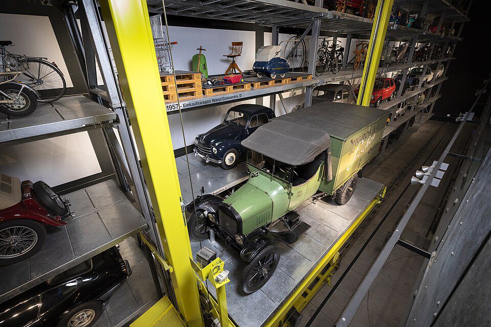 Interior de museu com estantes metálicas amarelas de múltiplos níveis exibindo veículos históricos, destacando um caminhão de entrega verde vintage e carros clássicos.